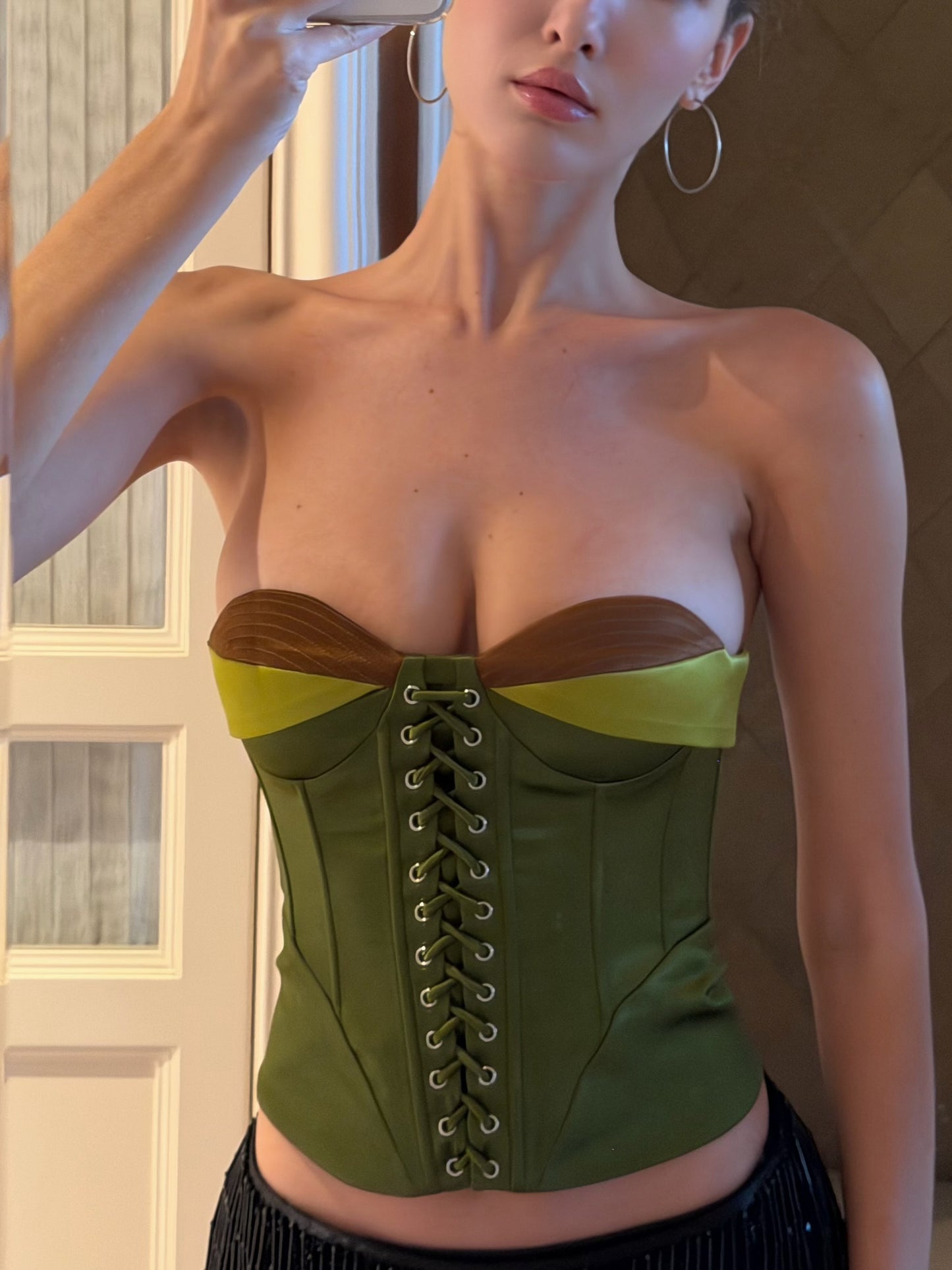 A woman wearing a structured strapless corset top in olive green with brown side panels and lime green underbust bands, shown in a mirror selfie.